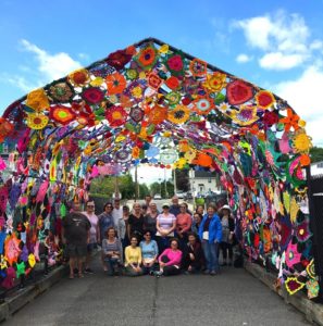 A group of 20 individuals posing under the tunnel of love, a colorful, crocheted flower dome, on a bright sunny day