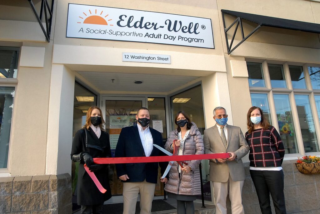 Five individuals standing outside of Elder Well Adult Day Care, cutting an opening ribbon on a sunny day