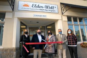 Five individuals standing outside of Elder Well Adult Day Care, cutting an opening ribbon on a sunny day