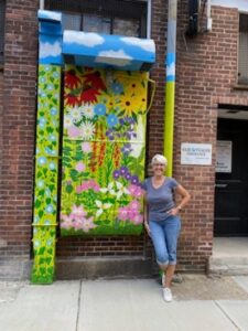 Artist Ginger McEachern in a grey t-shirt and jean capris, posing next to the public art display The UnconVENTional Garden, featuring a flower garden painted on metal