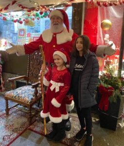 Santa Clause smiling with his arms out, standing with a young girl in a "Santa like" dress holding a wooden staff with sleigh bells, and another young girl in all black