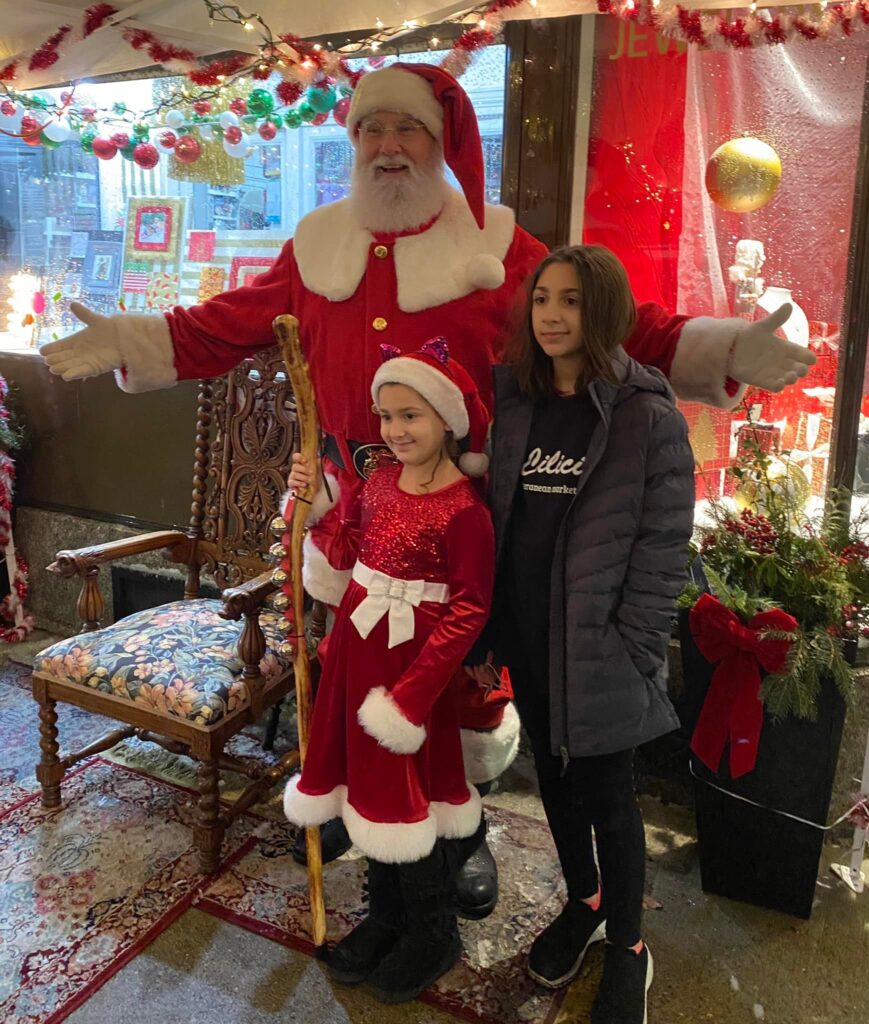 Santa Clause smiling with his arms out, standing with a young girl in a "Santa like" dress holding a wooden staff with sleigh bells, and another young girl in all black