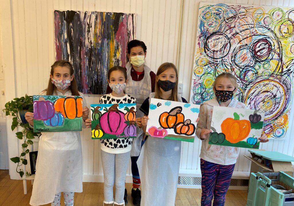 Artist Amy Adams standing behind four students who are holding up their vibrant pumpkin paintings, in a studio