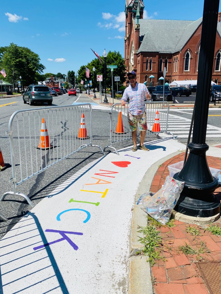 One of the artists of the Traffic Calming Project, wearing a grey shirt and khaki shorts, posing with his in-progress and colorful sidewalk art
