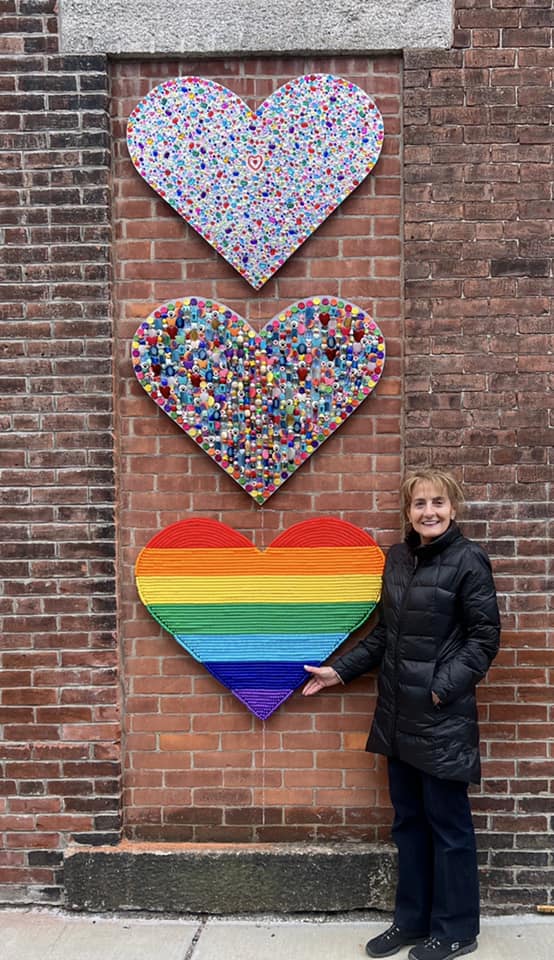Artist Jo LaFalce, in a black coat, standing next to her public art instillation The “Beading” Hearts on Adams Street in Natick Center.