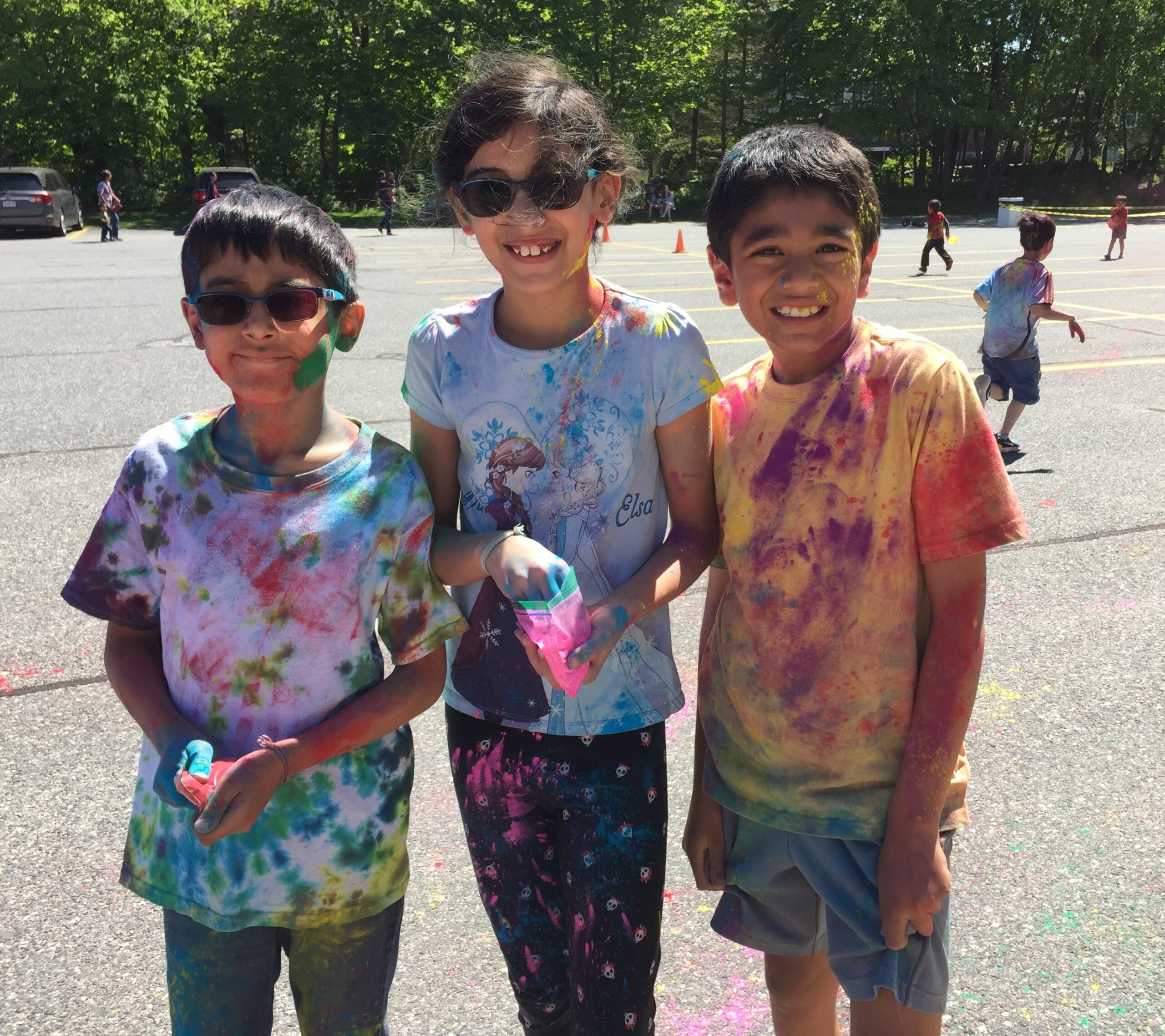 three children, (l-r) boy, girl, boy, wearing tie dye shirts, covered in colored chalk, smiling on a sunny day
