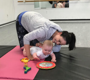 Woman in a grey shirt on her knees, bent over, playing with a happy baby during tummy time, on a pink yoga mat
