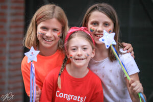 Three girls holding two star wands. The two on the Left and middle have on red shirts and the one on the right has a white shirt.