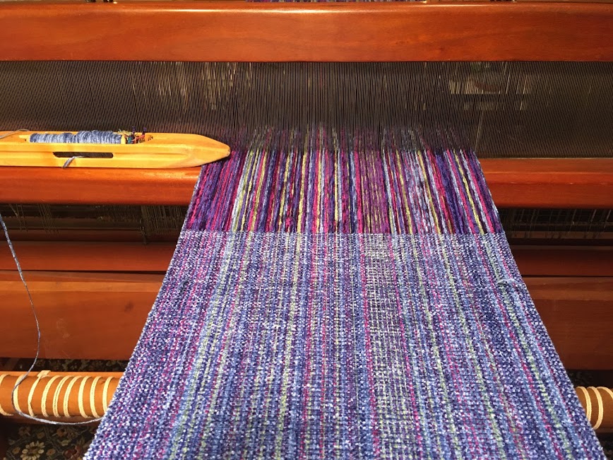 Color photograph of multicolor fabric strings being woven together on a wooden loom.