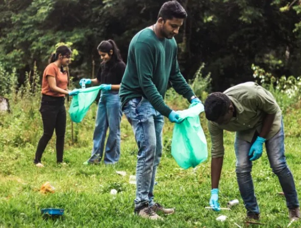 Modern color photograph of two adults and two teen girls picking up trash in a field with trees in the background.