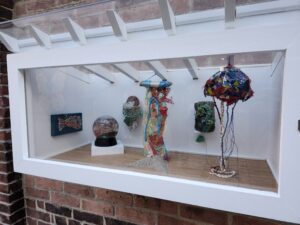 Modern color photograph of a miniature art gallery painted white with a wooden floor. In the gallery are ocean items made out of ocean debris.