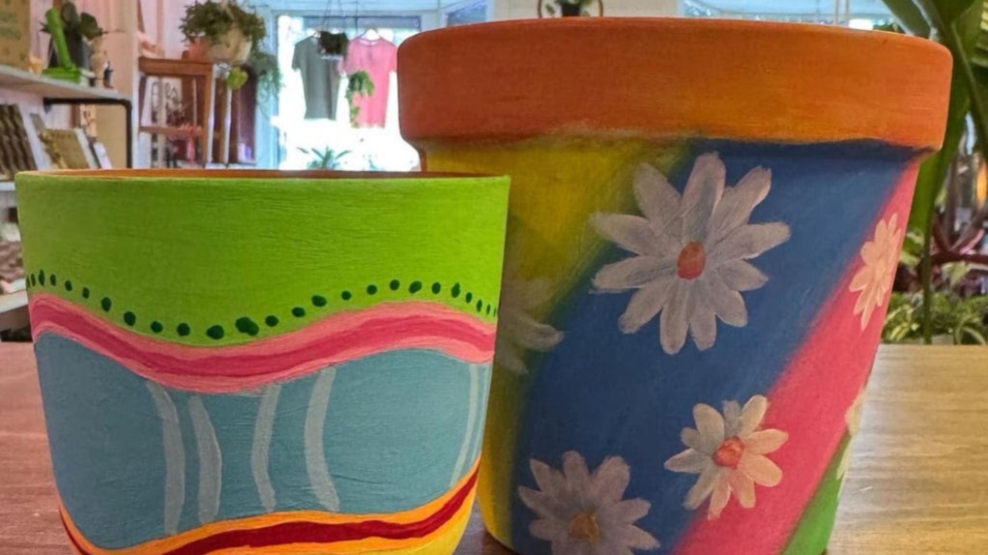 Modern color photograph of two painted clay plant pots. One is green, blue, pink, yellow and red in an abstract design, the other is blue, yellow and pink in angled stripes with white daisy flowers over the stripes.