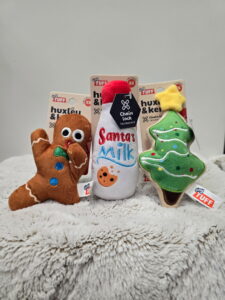 Modern color photograph of dogs toys. From left to right: a gasping gingerbread cookie doll, a glass milk bottle labeled "Santa's Milk," and a Christmas tree cookie doll.