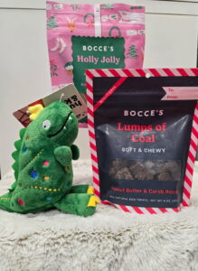 Modern color photograph of dog toys and treats. From left to right, a dinosaur doll, a pink package of treats, and a black package of treats labeled, "lumps of coal."