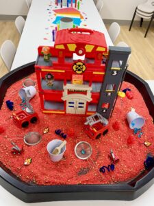 Modern color photograph of red play sand with many toys in it and a large red play firehouse with many floors, but it only goes back one room.