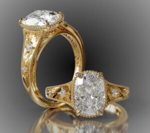 A modern color image of two views of the same gold and diamond ring on a gray background. One view is of the ring laying down, the stone pointed at the viewer, the other is of the side of the ring, the stone pointing up and slightly viewers left.