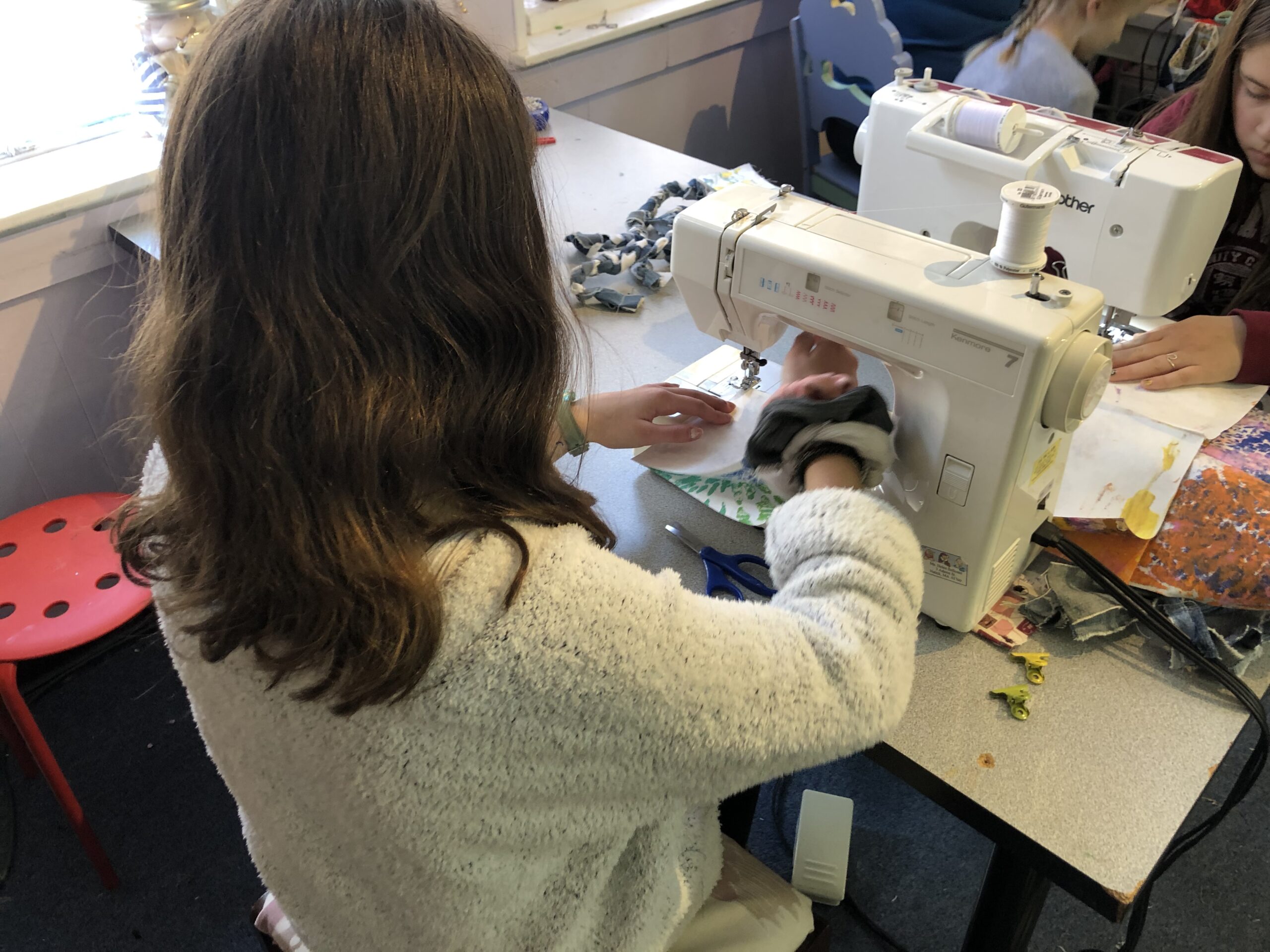 A modern color photograph of a girl with long brown hair, wearing a white sweater, working at a sewing machine. The view is over her right shoulder.