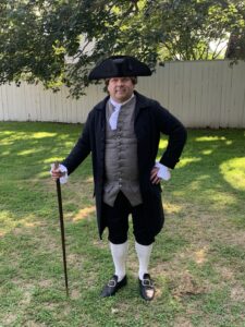 A modern color photograph of a middle-aged white man in 18th century costume. He stands on a grass lawn with a tan fence behind him. His costume consists of a long dark coat, tricorner hat, black pants to his knees, high cream socks to his knees, black shoes, a tan vest and a black cane. 