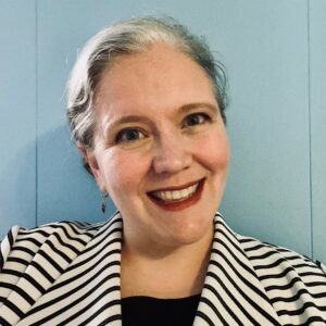 Modern color photograph of a middle-aged white woman with white hair and red lipstick. She is smiling and searing a black and shite striped jacket and a black shirt. the background is a light blue.