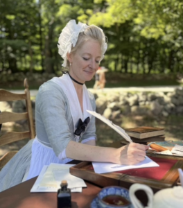 A modern photograph of a middle-aged white woman in an 18th century costume holding a quill pen to paper at a desk. The background is outside. 