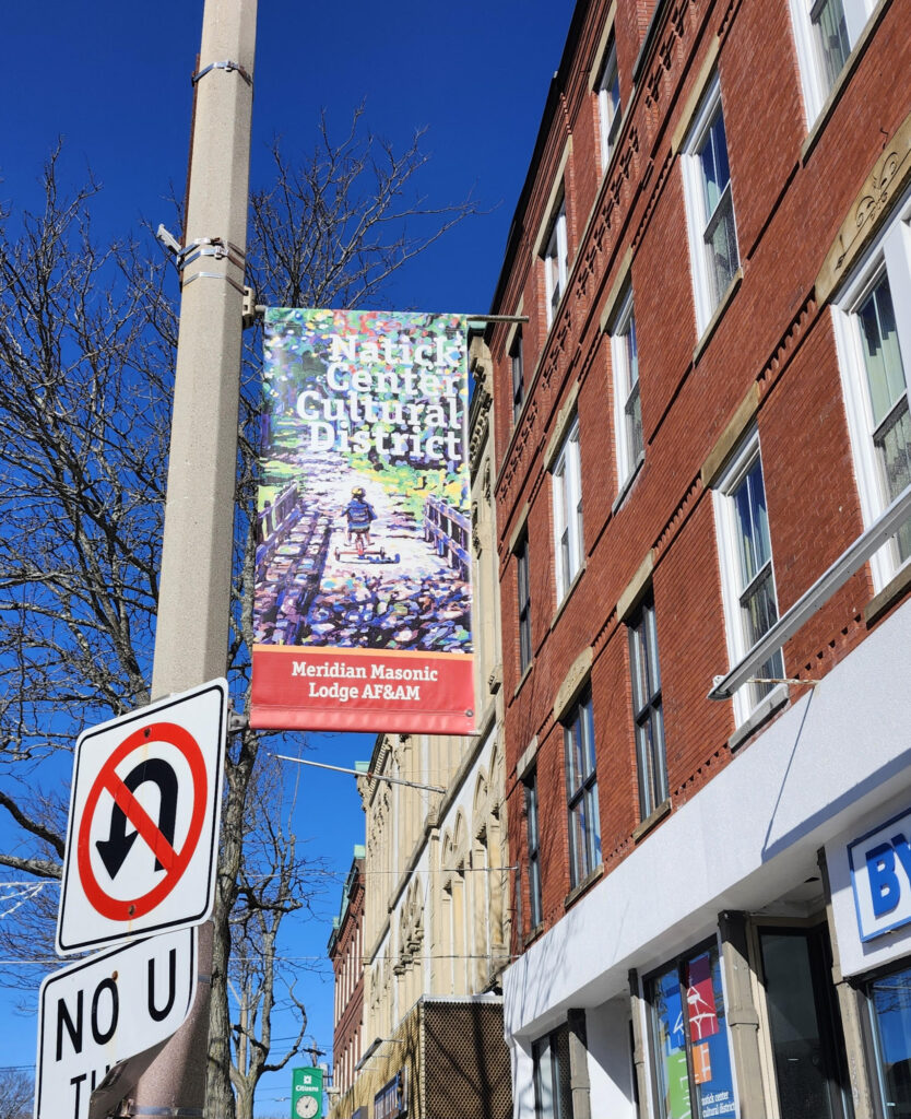 Modern color photograph focused on a street pole banner that has the name of a business and the Natick Center Cultural District name over a work of art of a child riding a bicycle. In the background is a clear blue sky and brick buildings.