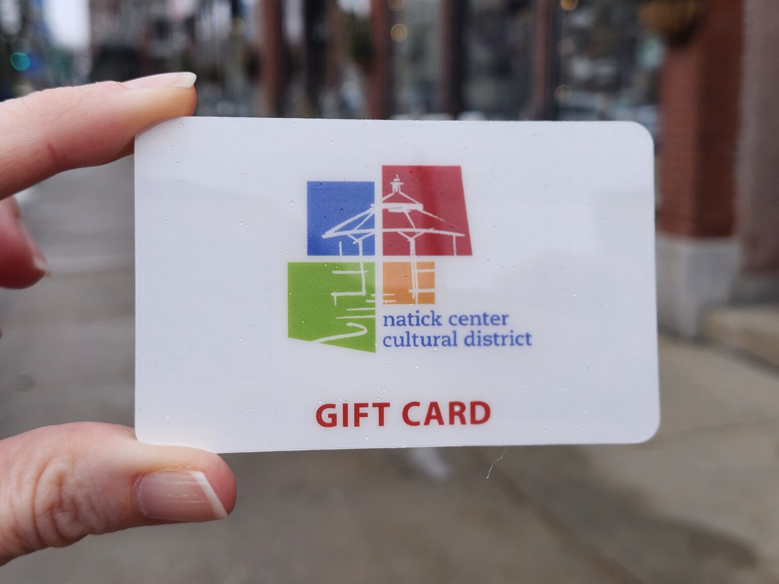Modern color photograph of a gift card with the colorful Natick Center Cultural District logo on it and red letters below it which reads, "Gift Card," in all capital letters. There are two fingers holding the card to the left and the background is a blurry streetscape.