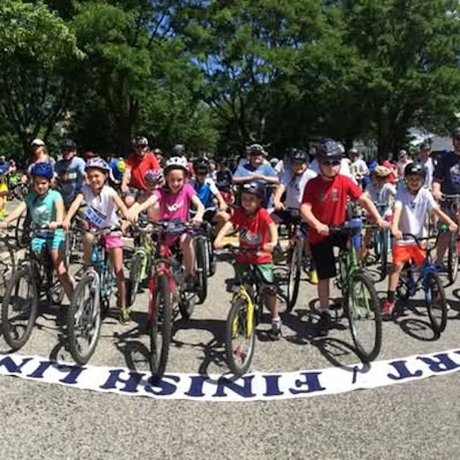 Modern color photograph of many kids and adults crossing the finish line riding on bicycles. All are wearing helmets and t-shirts. In the background are trees.