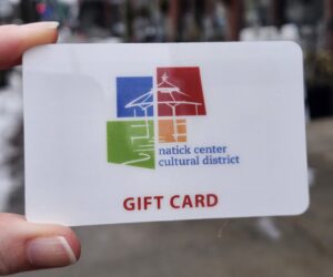 A modern color photograph of a white background gift card with a colorful logo for the Natick Center Cultural District. Red letters at the bottom read, "Gift Card."
