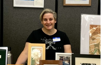 A modern color photograph focusing on a middle-aged white woman with short blonde hair who is smiling at the viewer and wearing a black shirt with a nametag on it. In front and behind her are framed and unframed pieces of art printed on paper.