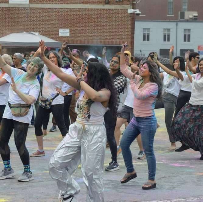 A large group of people of different races are dancing together in a group, many are wearing white with colored powder splashed on them. They are on pavement with a brick building behind them.
