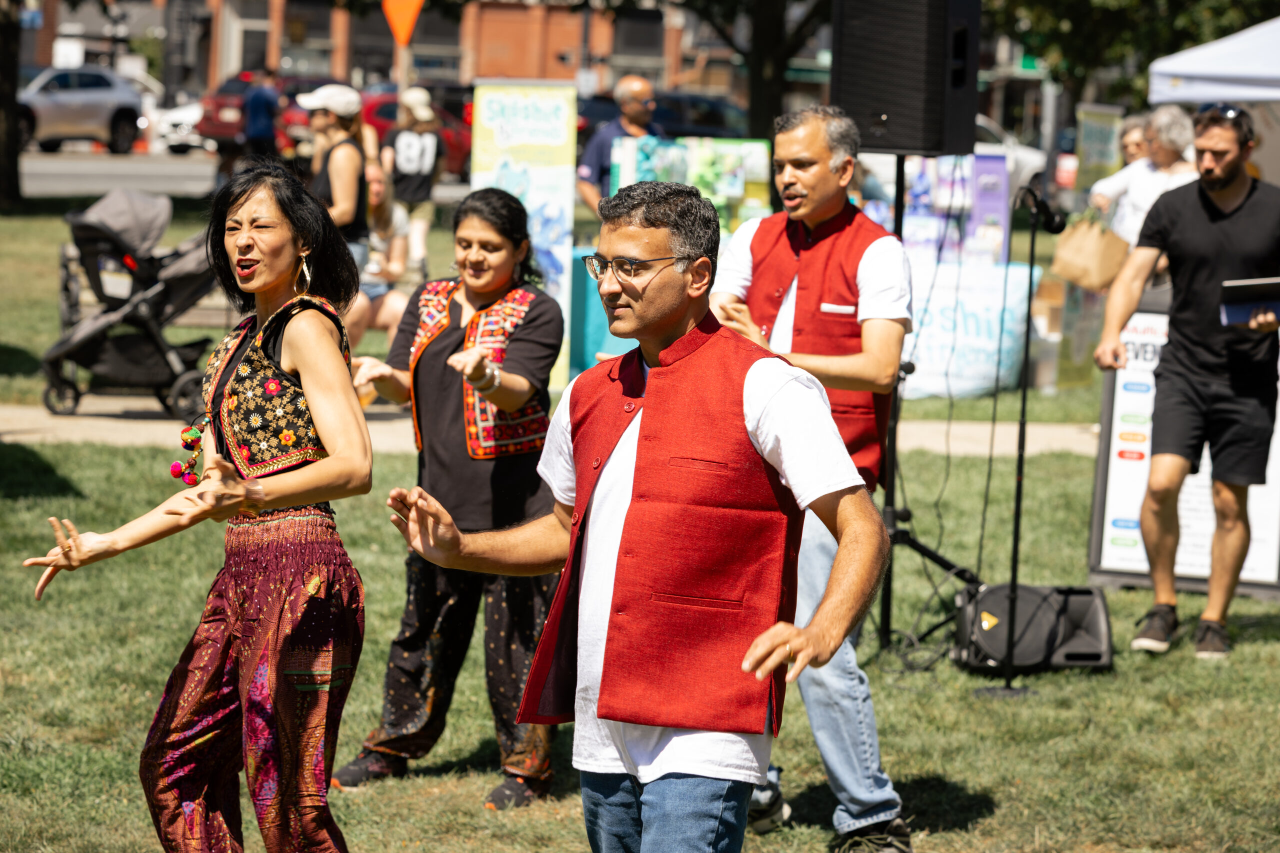 A color photograph of 4 adults dancing in loose costumes. The 2 men in red vests and white shirts with jeans, the women in not matching outfits, but loose fitting clothing in bright colors and black and brown. They are all on a grassy area with brick buildings behind them.