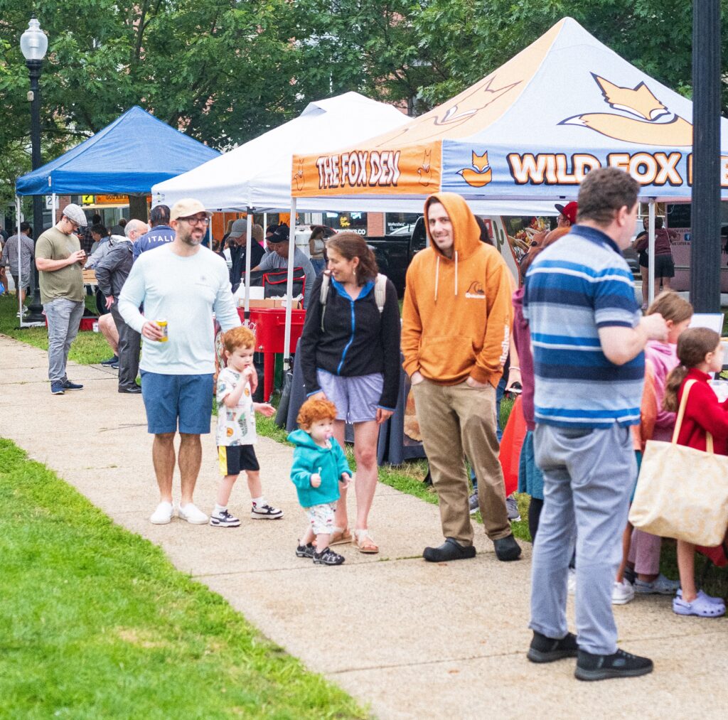 A modern color photograph of an outdoor scene. There are many people in street clothes who are walking up a paved sidewalk in a green grass park. Behind the people are tents.