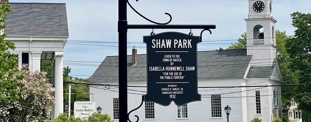A modern color photograph focusing on a black hanging sign for "Shaw Park," with a white steepled wooden church in the background, and a sunny, clear sky. Art in the Park