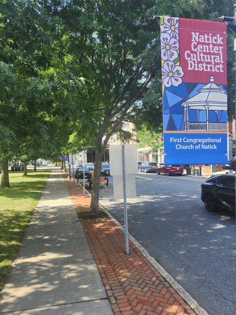 A modern color photograph of an urban scene at a park with trees lining the street, brick buildings on the other side and a banner on the street pole.