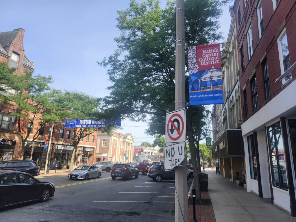 A modern color photograph of an urban scene with brick buildings, cars on the road and a banner over the street and on a street pole.