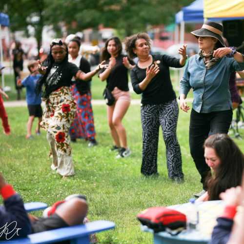 A modern color photograph of 5 feminine figures dancing, one Black woman in front of the other four of varying races. They are mostly dressed casually and are standing in a field with other people seated around them.
