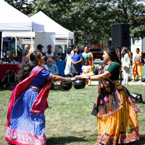 A modern color photograph of two Indian-American women in Saris holding hands and spinning. They are on a grassy field with tents and booths behind them. There are people in the background watching the dance.